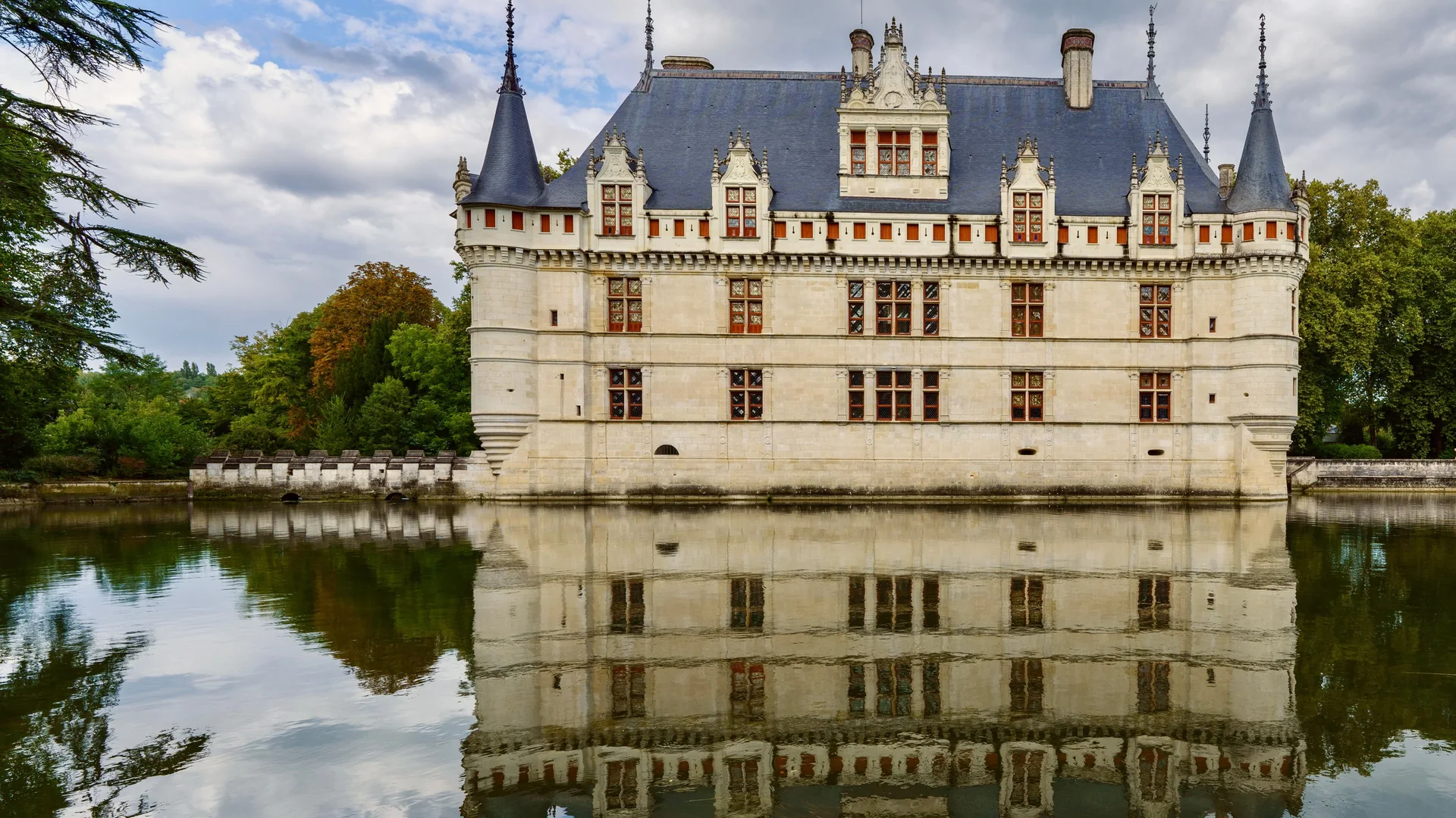 Bledě kamenná fasáda, břidlicové střechy a špičaté věžičky zámku Château d'Azay-le-Rideau se v klidné hladině řeky Indre odrážejí v celé své kráse, orámované stromy